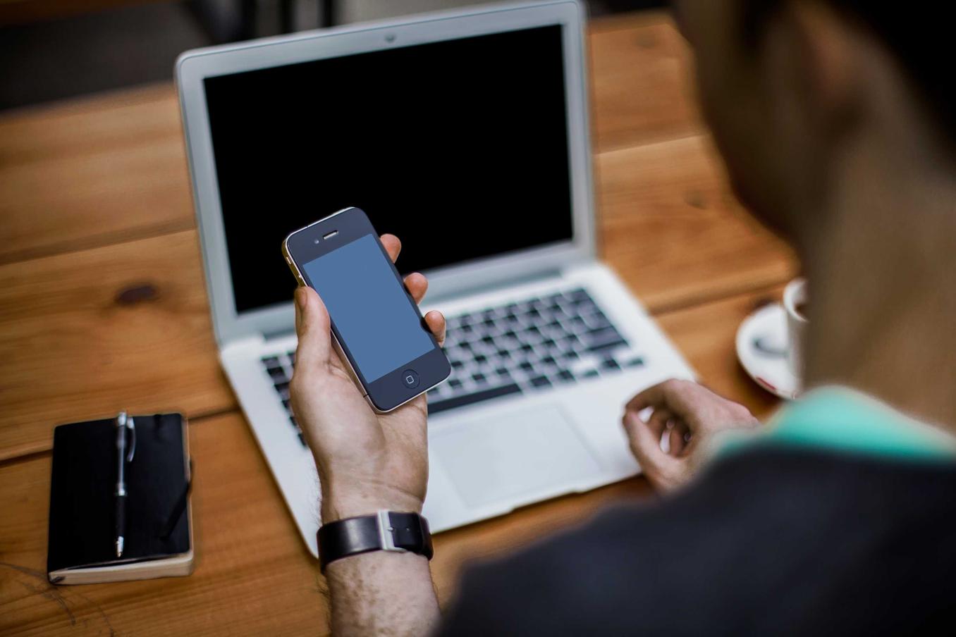 A man holding a phone and sitting in front of his laptop.